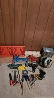 Photo showing the full contents of the lot including the vice, drill, soldering tool, hardware cabinet, work light, caulking tools, measuring tape, and various hand tools arranged on the floor against a brown wood paneled wall.