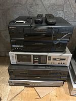 Stacked electronic devices consisting of Quasar and Sony VHS players, Aurex cassette deck, Technics CD changer, and Motorola HDTV receiver