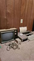 Vintage Zenith black and white television next to Commodore computers and accessories on floor against wood panel wall.