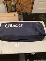 Dark blue Graco carry bag containing baby travel crib, shown laying flat on a white table indoors.
