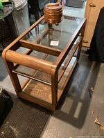 Diagonal front view of the wooden rolling bar cart with glass top showing its wooden frame, glass top and shelf, and small wooden barrel-shaped ice bucket on top.