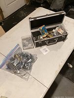 Photo showing hinges and hardware in a black case with some plastic containers and a clear plastic bag on a white table surface.