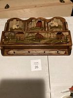 A front view photo of a wooden wall-mounted mail organizer with a hand-painted farm scene showing barns, fences, and sheep with three vertical compartments and a lower horizontal rack.