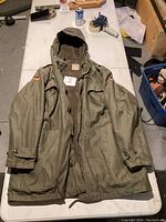 Front view of green military-style parka jacket with German flag patches and hood, laid out on a table.