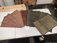 Ten wooden or bamboo style placemats arranged on a white table; four are reddish brown with lattice pattern, three are dark brown bamboo slats, and three have wider wooden plank designs in brown and greenish tones.
