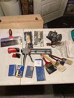 Photo showing entire assortment of tools including caulking gun, sanding blocks, paint brushes, putty knives, scraper, respirator mask, and a small red Milwaukee tool.