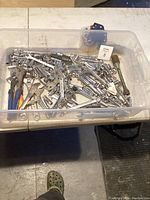 Clear plastic bin with assorted metal hand tools including pliers, socket wrenches, and various sockets.