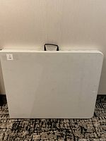 Folded white folding table standing upright showing handle on top and rounded edges.