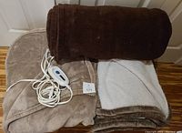 Three brown fleece blankets: one electric with controller and cord, one dark brown rolled, one light brown with white inner lining, displayed on wooden floor.
