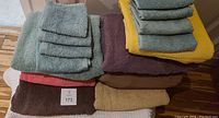 Photo showing a collection of full-size towels in multiple colors including green, yellow, brown, red, burgundy, and beige. Each towel is paired with matching washcloths stacked on top.
