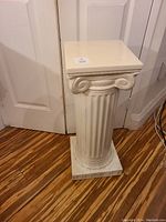 Off-white Greek style ceramic pedestal with glossy finish, standing 26 inches tall, shown against a door and wooden floor.
