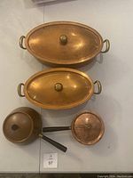 Top view of copper pots and pans with lids laid out, showing oval pots with lids and small saucepans with black handles.