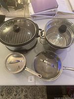 Top-down view of various stainless steel pots and pans including a frying pan with glass lid and a pot with steamer insert