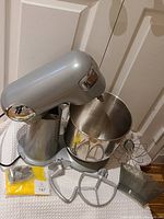 Photo of a silver Cuisinart stand mixer with stainless steel mixing bowl. Included accessories are the flat beater, dough hook, whisk attachment, and a yellow icing bag containing several metal piping nozzles.