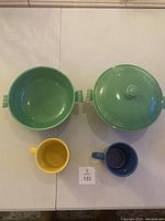 Overhead view of two green Fiesta bowls (one with lid with chips) and two mugs (yellow and blue) on white surface.