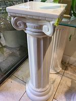 Photo of an off white glossy Greek style pedestal with Ionic column design showing fluted shaft and scroll capitals