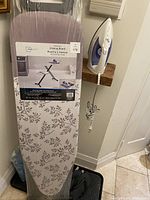 Photo showing full ironing board with packaging and floral cover, alongside the iron hanging on a wall mount.