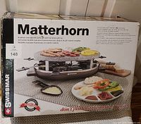 Photo of unopened boxed Matterhorn oval raclette party grill with hot stone top, showing packaging and product image with 8 raclette pans.