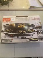 Box front showing the Trudeau Maison 18-piece raclette party grill set with a photo of the product in use and branding details.