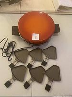 Tefal orange dome-shaped raclette grill with six triangular pans arranged in front, showing the appliance and accessories