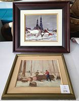 Photo showing two framed winter-themed artworks; one with church steeple and snowy village scene, the other with lumberjacks working around logs in snowy forest.