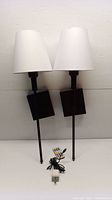 Two black metal wall lamps with square base mounts and long stems, topped with white fabric shades. Shown off and unlit. Included remote and power adapter visible on bottom.