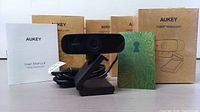 One AUKEY 1080P Webcam out of the box showing the webcam body with lens, clip mount, USB cable, user manual, and reference card.