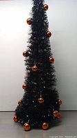 Full view of the tall, slim black tinsel Christmas tree decorated with shiny orange spherical ornaments.