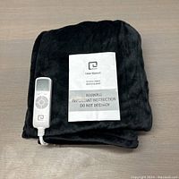 Photo of folded black heated throw blanket with wired remote controller and user manual on top on a table surface.