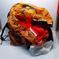 Orange inflatable dinosaur costume laid down, showing open mouth with red interior and white teeth, black spots on orange material, resembling T-Rex