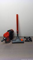 Vacuum cleaner main unit with orange extension tube and two attachments on a wooden surface against a plain wall.