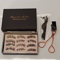 Open black box showing 12 magnetic eyelashes arranged in pairs labeled for left and right eyes, top and bottom lashes. Magnetic strips visible on lash bands. Also shown black plastic applicator tweezer and rose gold eyelash curler.
