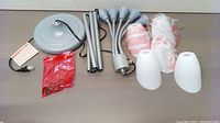 Lamp parts spread out: gray base, metal rods, adjustable arms, multiple white shades, power cord, assembly instructions and accessories in plastic bag.
