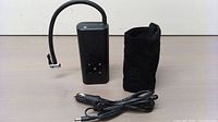 Portable black electric air pump with flexible inflation tube, power cord, and black soft pouch next to it.