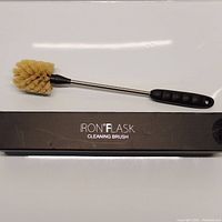 Side view of the Iron Flask Cleaning Brush lying on its box, showing the full length brush with black handle, metal shaft and tan bristles.