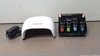 Photo of the MEFA UV LED nail lamp, power cord, and six acrylic gel paint tubes on a transparent stand on wood surface.