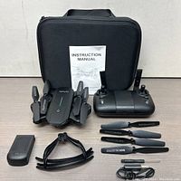 Drone and all accessories laid out including remote control, instruction manual, propeller guards, spare propellers, battery, USB charging cable, and black carrying case.