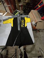 Front view of adult medium wet suit laid flat showing black, yellow, and gray sections with zipper closed halfway and RUA logo on chest.