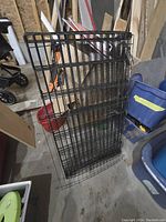 Black metal dog pen leaning against a wall in a storage area or garage with various items around.