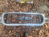 Image shows the front view of the GMC grill with chrome outer frame and black horizontal slats, placed on a metal stand with leaves on ground.