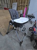 Side angle view of the Baby Trend kids high chair showing the gray padded seat, white tray with cup holder, and foldable metal legs in a storage or garage setting.