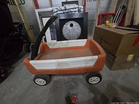 Orange and white Little Tikes kids wagon with black handle and wheels, shown indoors on concrete floor next to boxes.