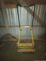 Manual lawn mower, yellow frame with black handle, against a metal wall background.
