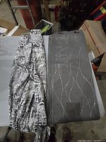 Four curtains displayed folded on a flat surface; two grey ones each with silver wave pattern and two white ones with black and grey floral patterns.