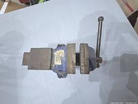 Top view of a small metal bench vise showing the blue painted body, metal jaws, and handle for tightening.