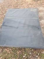 Full view of folded black tonneau cover lying flat on ground with some dirt spots visible.