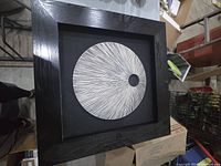 Shadow box wall decor showing a black frame with a black background and a white circular abstract design with radiating black lines and a black center dot.