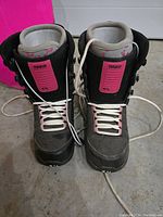 Front view of pair of women's snowboarding boots with black outer material, pink accent labels on tongue, white laces, gray padded lining.