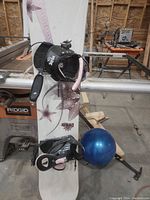 Full view of the snowboard standing against a workbench in a workshop, showing the top surface graphics and attached bindings.