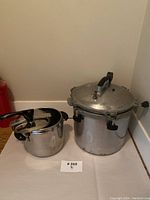 Two pressure cookers side by side on a white surface. The larger aluminum cooker shows wear and simpler handles, the smaller stainless steel cooker is shiny with black handles.